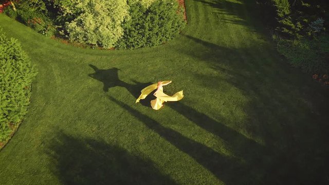 Joyful blonde woman in a bright yellow dress with a train, having fun and spinning in the park. Background green lawn in royal garden. The fabric flies, flutters in the wind. Establishing Aerial View.
