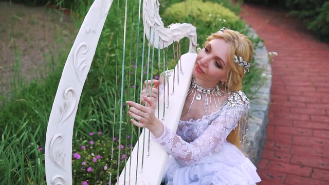 Attractive blonde woman in a white, creative, lace vintage dress plays the harp. Background fabulous royal garden. Holds an old musical instrument in his hands. Outfit, image of a gentle elf musician.
