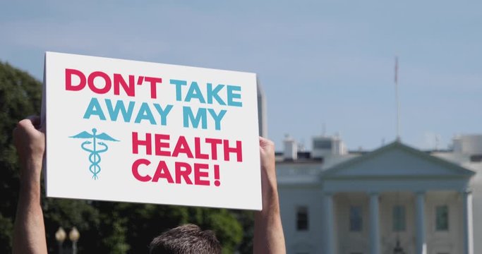 A man holds a DON'T TAKE AWAY MY HEALTH CARE protest sign in front of the White House on a sunny summer day.	