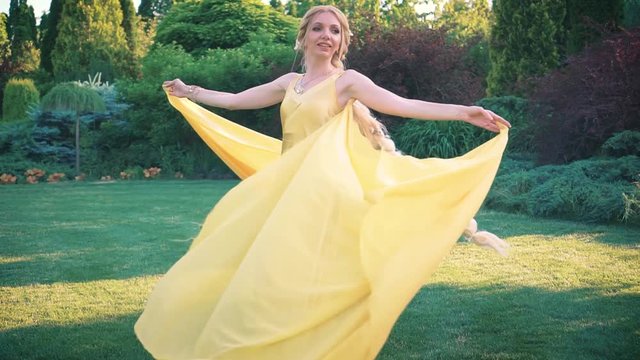 Joyful blonde woman with a long braid in a yellow dress. Cape train flies flutters in the wind. Rapunzel having fun is going round and spinning in the park. Background green trees landscaping lawn.