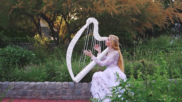 Attractive blonde woman in a white creative lace vintage dress plays the harp. Background fabulous royal garden. Stylish braided hairstyle with a silver diadem. Outfit, image of a gentle elf musician.