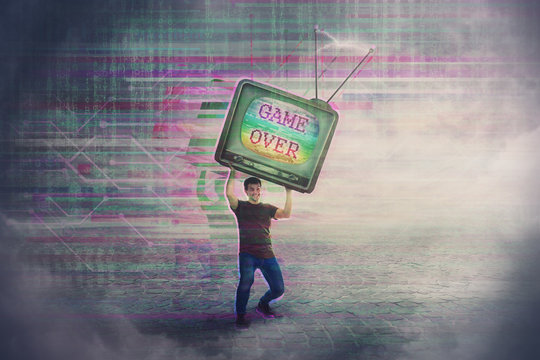 Angry gamer lifting up and throwing his old TV box after glitch text game over on the screen. Addicted, guy playing video games, lost in virtual reality world. Modern entertainment technology.