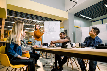Young woman standing near the white office board with chart and speaking to her colleagues . Multiracial team of people having meeting, testing virtual reality glasses in office sitting at the table