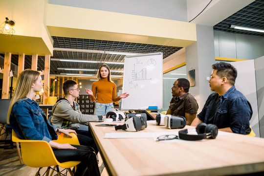 Multiethnical Business People Having Team Training Exercise During Seminar With VR Glasses. Young Girl In Orange Sweater Presenting Results Of The Project For Coworkers