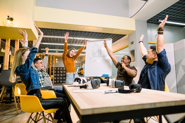 Young business team with hands up for new startup with vr goggles or victory in creative office. Cheerful diverse people giving strength motivation. Co-working and teamwork concept