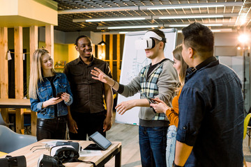 Business team of multiracial people working on virtual reality applications and games, young excited man testing VR glasses or goggles standing in the office room with colleagues