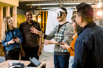Business team of multiracial people working on virtual reality applications and games, young excited man testing VR glasses or goggles standing in the office room with colleagues