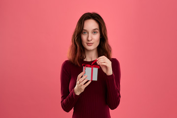 Excited pretty brunette girl opens gift packed in present box with a ribbon standing isolated on a dark pink background and smiling at the camera. Celebrating special event.