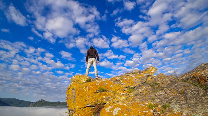 Man stands confidently on top. Blue sky with clouds, fog, mountains. View from the bottom and rear. Concept of achievement, success.