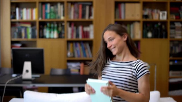 Portrait of pretty beautiful young woman dancing happily at home near a bookshelf. Modern student girl dancing throwing a book in modern house living room. End of school, vacations.