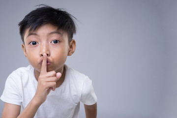 An Asian child wearing white t-shirt putting a finger on his lips a sign of quiet please on grey background. 