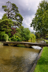 Sun on Pine tree and footbridge over the River Windrush after rain storm in Bourton-on-the-Water village in the Cotswolds England