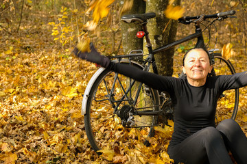 Woman in black clothes 58 years old with a bicycle on a background of fallen leaves. The woman sits and joyfully tosses up the yellow leaves. Copy space.