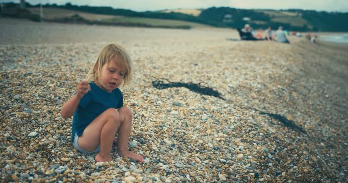 Little toddler sitting on the beach throwing stones