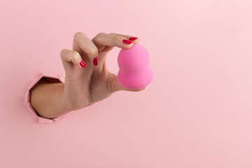 Girl hand holds a beauty blender for makeup from a hole in a pink background, copy space.
