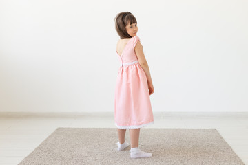 The little charming girl in a pink dress smiles and looks into the camera against the background of a white wall. Concept of the author's children's clothing. Copy space