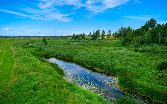 A narrow water canal, river, stream going through a green grass field landscape into bright summer clouds. Rural, countryside landscape