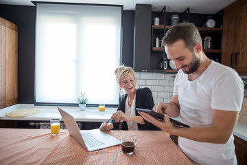 Obraz premium Couple in kitchen in the morning. They are smiling and browsing tablet and laptop.