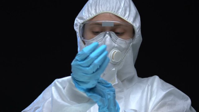 Scientist in protective suit glasses and respirator putting on gloves experiment