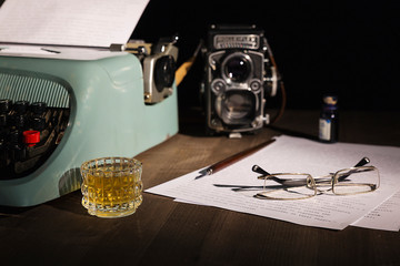 Vintage photograph of a journalist's office, with typewriter, fountain pen with inkwell, old bioptic camera and glass of whiskey.