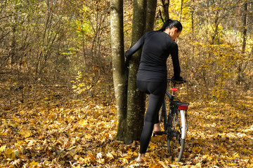 Barefoot woman of 58 years with black hair and in black clothes standing near bicycle behind on yellow autumn background in forest.