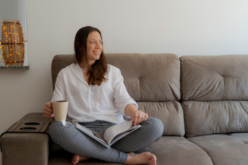 Beautiful smiling woman sitting on the couch reading magazine and having a tea.