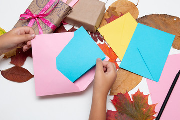 children's hand puts a card in an envelope, gifts with beautiful ribbons, greetings for the holiday, Thanksgiving, birthday, autumn leaves on a white table, copy space top view, pink, yellow, blue