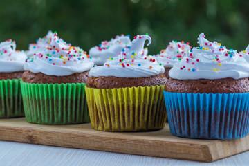 Delicious muffins with white cream and colored sprinkles on a wooden table on a background of green foliage.