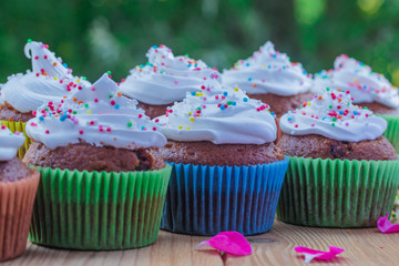 Delicious muffins with white cream and colored sprinkles on a wooden table on a background of green foliage.