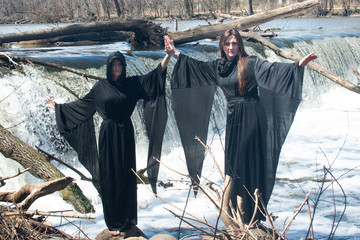 Two Women in black robes holding hands in front of a Waterfall. Witches. Halloween concept. Witchcraft and magic. 