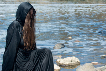 Hooded Woman with dark long hair in black robes in front of the Lake. Witches. Halloween and Gothic. Witchcraft and magic. Back to Nature concept.