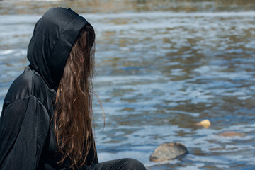 Hooded Woman with dark long hair in black robes in front of the Lake. Witches. Halloween and Gothic. Witchcraft and magic. Back to Nature concept.