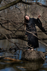 Woman with dark long hair in black robes standing on a tree stump in the middle of a swamp. Halloween and Gothic concept. Witchcraft and magic. Witch.