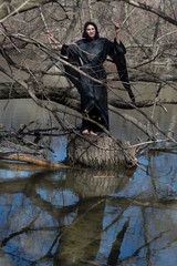 Hooded woman in black robes standing on a tree stump in the middle of a swamp. Halloween and Gothic concept. Witchcraft and magic. Witches.