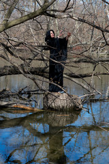 Hooded woman in black robes standing on a tree stump in the middle of a swamp. Halloween and Gothic concept. Witchcraft and magic. Witches.