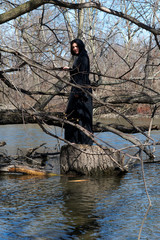 Woman with dark long hair in black robes standing on a tree stump in the middle of a swamp. Halloween and Gothic concept. Witchcraft and magic. Witch.