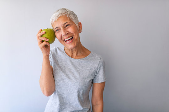 Portrait of a beautiful elderly woman holding an apple, smiling, isolated on gray background. Happy Woman Holding Granny Smith Apple. An aple a day keeps doctor away