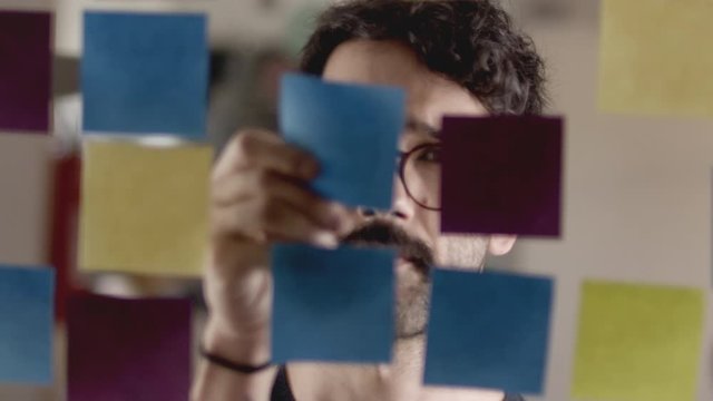 Close up of a young man brainstorming and placing a sticky note on a glass whiteboard.