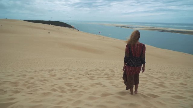 Famous Dune du Pilat near Bordeaux, France. A large sand dune by the atlantic seaside, filmed in slow motion. A young  brunette woman in a beautiful dress is looking at the beautiful nature.