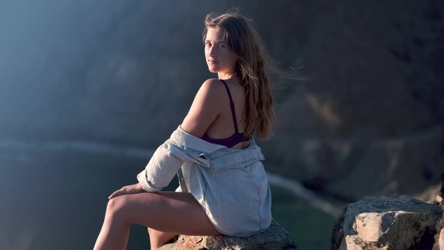 Slow motion of a young blond girl sitting on a rocky cliff, with the sea in the background, on a windy evening