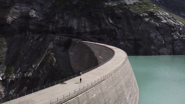 Aerial view of the Limmernsee reservoir and dam near Linthal (Switzerland) while someone is walking on the dam. Orbital drone flight above the dam. The cliffs of the alps can be seen in the background