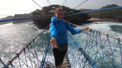 PoV action camera - funny woman invite to go on suspension bridge above ocean waves in Timang beach, Java island, Indonesia