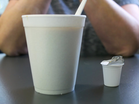 Styrofoam coffee cup with plastic spoon and used creamer container in foreground and human elbows on table in background.