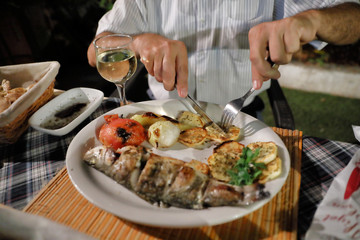 Dinner at the restaurant. Mediterranean food. Grilled fish and vegetables on a plate. Hands of a man cutting fish in a plate.