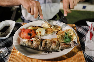 Delicious Dinner at the restaurant. Mediterranean food. Grilled fish and vegetables on a plate. Hands of person cutting fish in a plate. Top wiev