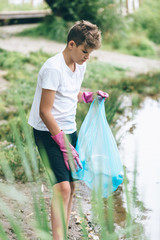 Boy in white t shirt in gloves collects garbage and plastic bottles into blue package on the beach. Young volunteer. Environmental protection, save environment concept. Close up hands in gloves