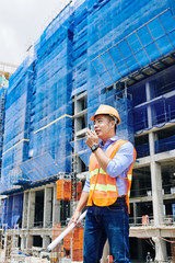 Young Vietnamese head engineer talking on walkie-talkie and giving orders to workers at construction site