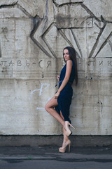 A glamorous woman in a short evening dress with heels stands next to the concrete wall.