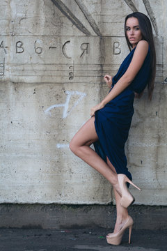 A glamorous woman in a short evening dress with heels stands next to the concrete wall.
