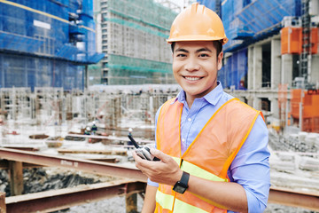 Portrait of cheerful Vietnamese construction engineer standing with portable radio communicator and...